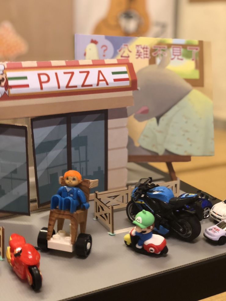 孩子們七手八腳地，大家合力把汽機車模型都移開，擺放好，讓出一條通路來。 並讓孩子們輪流幫忙推著輪椅公仔玩偶進披薩店買披薩。